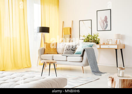 Blanket and pillows on a sofa and table on a carpet against a wall with posters in cozy living room with mattress Stock Photo