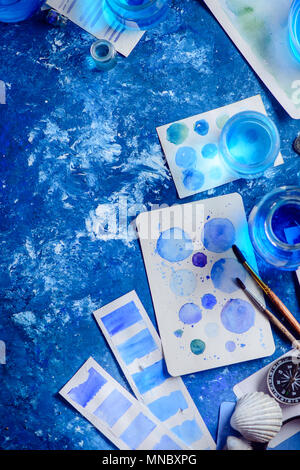 Blue shades artist workplace with sea watercolor, palettes, sketches, brushes and water jars, seashells and compass. Painting sea concept on a navy bl Stock Photo