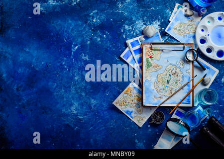 Fantasy maps on a wooden clipboard in an artist workplace with sketches, paint, brushes. Travel essentials flat lay with binoculars and compass on a n Stock Photo