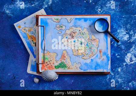 Sea travel and painting concept. Watercolor fantasy map on a wooden clipboard with a magnifying glass on a navy blue background with copy space. Stock Photo