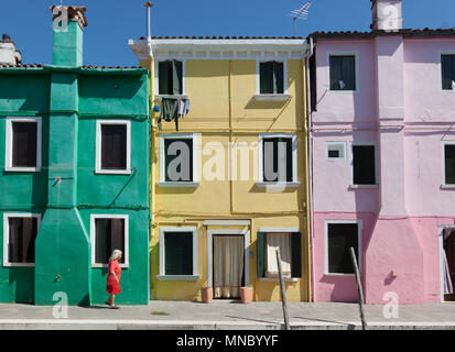 View of Senior woman with colorful dress walking across colorful facade in the island of Burano near Venice - Italy Stock Photo