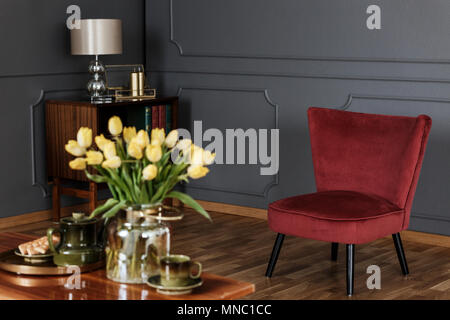 Red armchair in dark grey living room interior with lamp and yellow flowers on table in blurred foreground Stock Photo