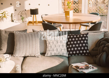 Black and white decorative pillows on a gray sofa in a sunlit living room interior with a wooden dining table Stock Photo