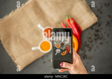 Focus on a woman's hand holding a black phone and using it to take a photo of an arrangement of spices and red and yellow peppers Stock Photo
