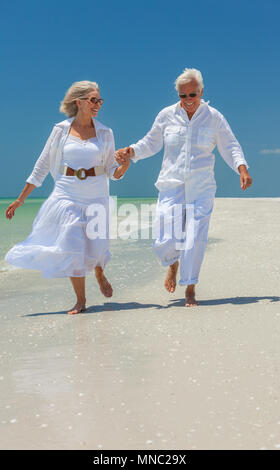 Happy senior man and woman retired couple wearing sunglasses holding hands and running on a deserted tropical beach with clear blue sky Stock Photo