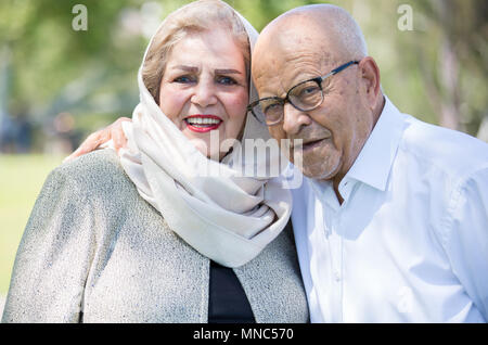 Closeup portrait, retired couple in white shirt and dress holding each other smiling,enjoying life together, isolated outside green trees background. Stock Photo