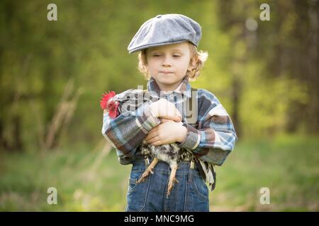 Small boy portrait with chicken. Caucasian curly haired boy in hat holding small chicken in outdoor. Stock Photo