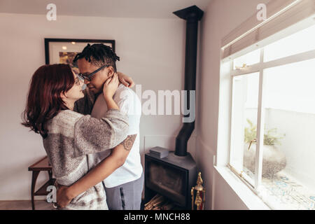 Beautiful multi-ethnic couple looking each other with love while standing indoors near window. Romantic man and woman embracing each other at home. Stock Photo