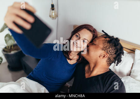 Romantic multiracial couple lying together on bed and taking selfie. Smiling man kissing on the cheek of woman taking selfie with mobile phone. Stock Photo