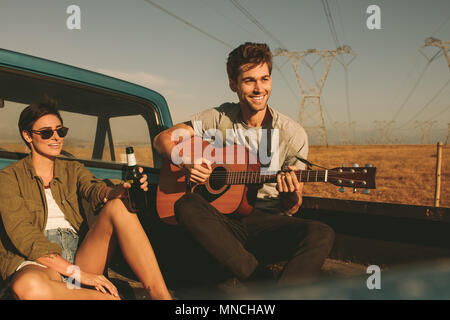 Couple sitting in the back of their pickup car enjoying the road trip in country side. Man playing the guitar while the woman is holding a bottle of d Stock Photo