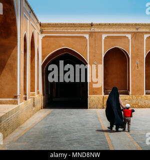 Unidentifiable woman wearing a traditional black dress in Iran, known as a chador, holds the hand of a young boy. Islamic architecture in background with ancient arches Stock Photo