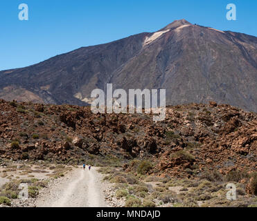 Two walkers lend scale to a lava flow and the distant summit cone of Mount Teide or an active volcano on Tenerife in the Canary Islands Stock Photo