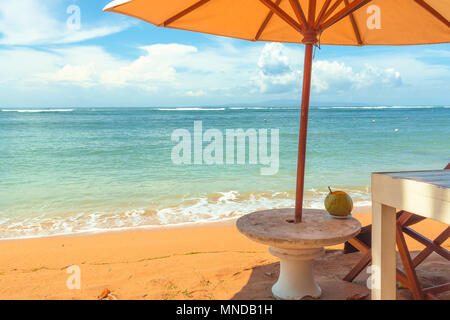 A chair and a stone table on the yellow beach under a parasol, relaxing with a coconut-drink. Sanur, Bali, April 21, 2018 Stock Photo