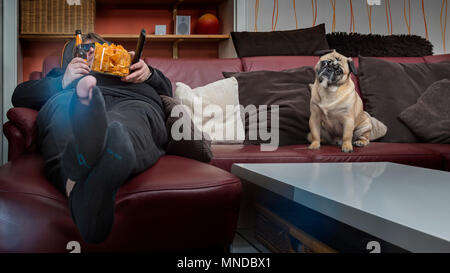 A man eats chips in front of the TV. His dog is sitting next to him on the couch. The man has a hole in the stockings and a 3 D glasses put on. Frog p Stock Photo