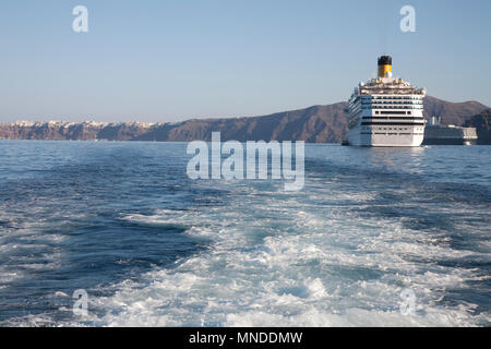 The water behind a large boat trails into the ocean, with two large cruise liners in the background and a village Stock Photo