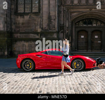 Royal Mile, Edinburgh, 16th May 2018. Tourists enjoying the sunshine on the Royal Mile, Edinburgh, Scotland, United Kingdom. A young woman walks looking at a phone carrying a Dolce and Gabbana handbag past a bright red Ferrari 488 GTB coupe sports car parked on a double yellow line on the cobbled Royal Mile next to St Giles Cathedral Stock Photo