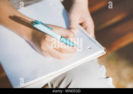 Close up view on boy's hands writing in notebook outdoor. Teenager sitting on a wooden bench and doing his homework. Stock Photo