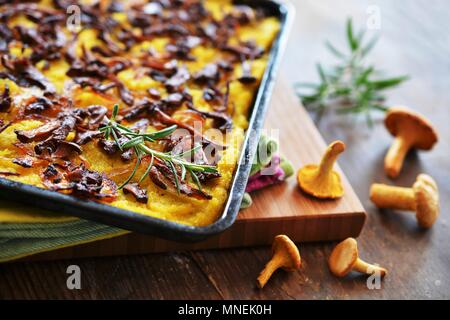 Polenta cake with chanterelle mushrooms and onions on a baking tray next to fresh mushrooms and sprigs of rosemary Stock Photo