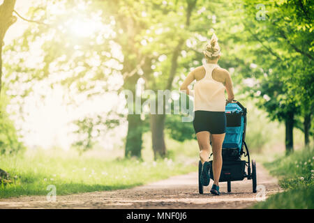 Running woman with baby stroller enjoying summer day in park. Jogging or power walking supermom, active family with baby jogger. Stock Photo