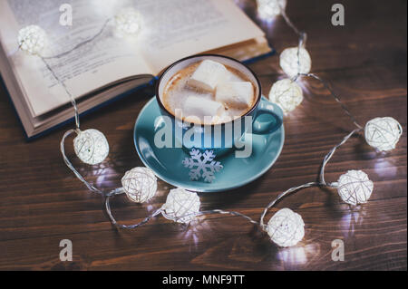 Cozy winter home. Cup of cocoa with marshmallows, open book, Christmas garland, on a wooden table. Kind atmosphere of evening  reading.