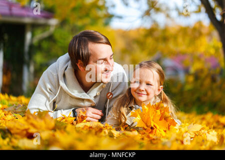 Father and his adorable little daughter outdoors on sunny autumn day laying on ground covered with fallen yellow leaves Stock Photo