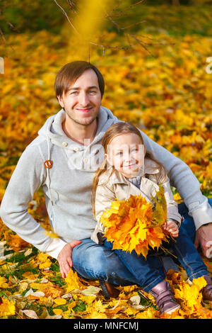 Father and his adorable little daughter outdoors on sunny autumn day laying on ground covered with fallen yellow leaves Stock Photo