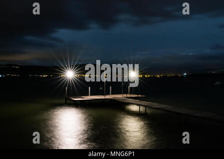 swimming pier on a lake at night with two lights burning bright over shimmering water and the lights on the opposite shore glowing in the distance Stock Photo