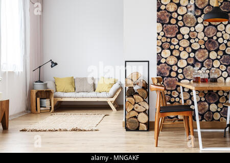Natural design in open plan studio with log holder, wood wallpaper in separated dining room and wooden box standing next to sofa in living space Stock Photo