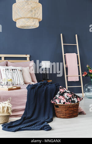 Decorative floral cushion in brown basket next to bed with blue blanket Stock Photo