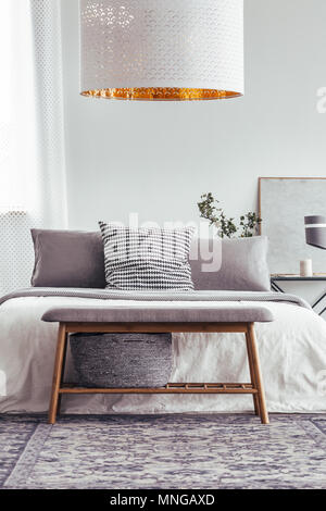 Designer white lamp above wooden bench on patterned carpet in bedroom with grey pillows on bed Stock Photo