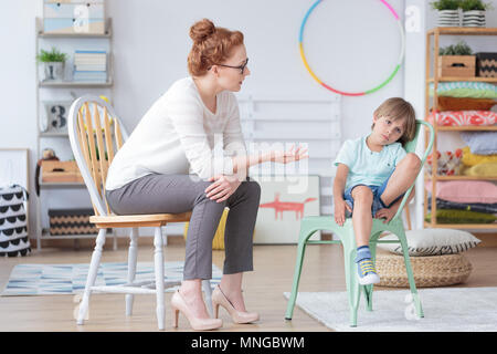 Red haired counselor talking with worried boy sitting on mint chair in colorful room with toys Stock Photo