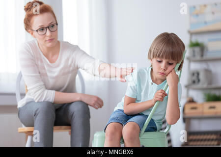 Sad autistic boy sitting on a mint chair during session with red haired psychotherapist Stock Photo