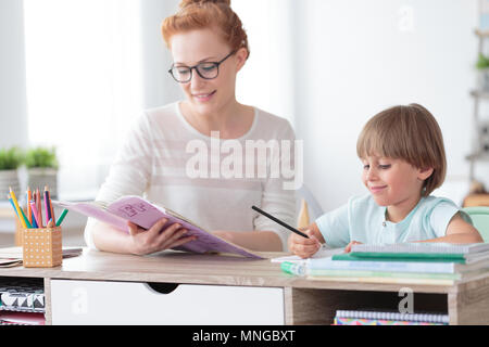 Math teacher doing exercises from workbook with young happy boy while sitting at desk in classroom Stock Photo
