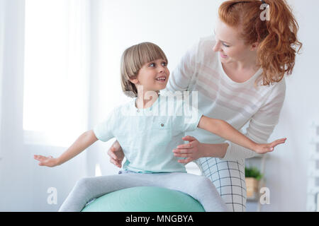 Red haired physiotherapist supporting boy with scoliosis doing correction exercise on mint ball Stock Photo