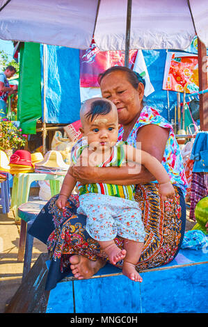 KYAIKTIYO, MYANMAR - FEBRUARY 16, 2018: The elderly Burmese woman is holding her grandson on the knees, sitting under the sunshade in market street of Stock Photo