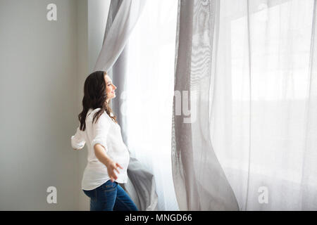 Happy pregnant woman opening window curtains Stock Photo