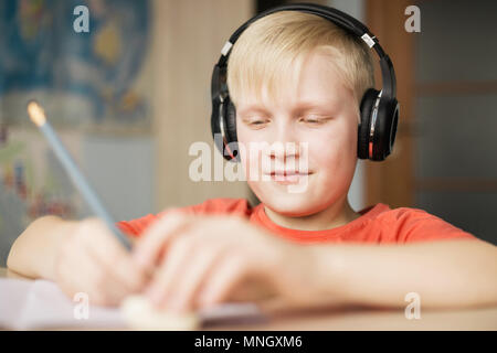 Blond caucasian boy in headphones writing and smiling, doing his homework. Stock Photo