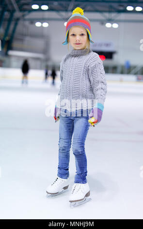 Adorable little girl wearing jeans, warm sweater and colorful hat skating on ice rink Stock Photo