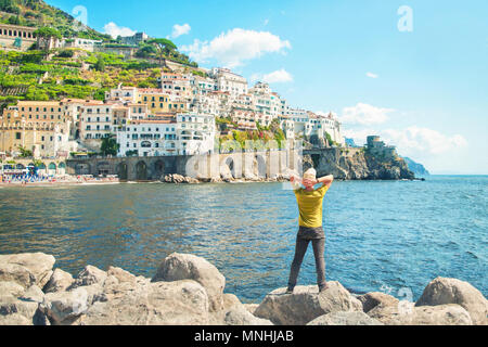 young caucasian man standing on rocks by sea with his back to camera enjoying view of small european coastal town Stock Photo