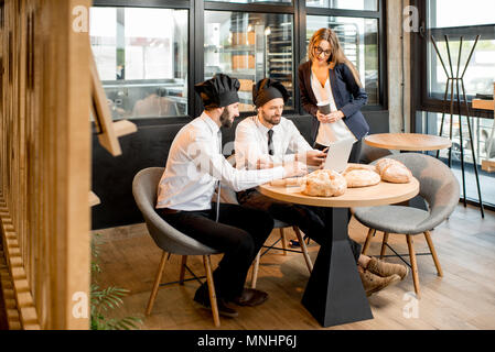 Businesswoman with two elegant bakers or chef cooks working with laptop in the cafe with kitchen on the background Stock Photo