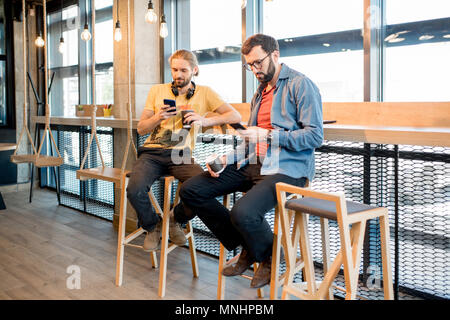 Two men dressed casually using smartphones sitting near the window of the modern cafe interior Stock Photo