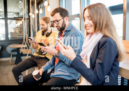 Young people dressed casually using smartphones sitting near the window of the modern cafe interior Stock Photo