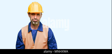 Senior engineer man, construction worker with sleepy expression, being overworked and tired isolated over blue background Stock Photo