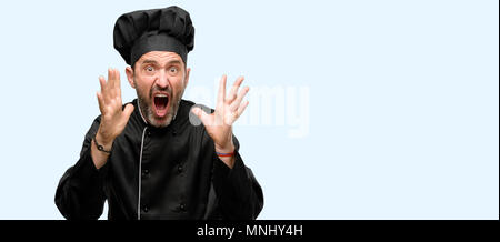 Senior cook man, wearing chef hat stressful keeping hands on head, terrified in panic, shouting isolated over blue background Stock Photo