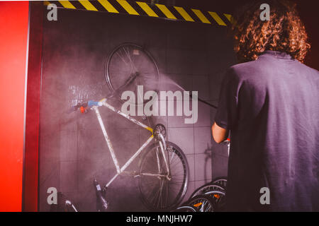 Services professional washing of a bicycle in the workshop. Close-up of hand Young Caucasian stylish man doing bicycle cleaning using automatic electric water pump. Sprays scatter from the pressure Stock Photo