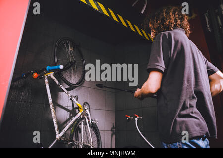 Services professional washing of a bicycle in the workshop. Close-up of hand Young Caucasian stylish man doing bicycle cleaning using automatic electric water pump. Sprays scatter from the pressure Stock Photo