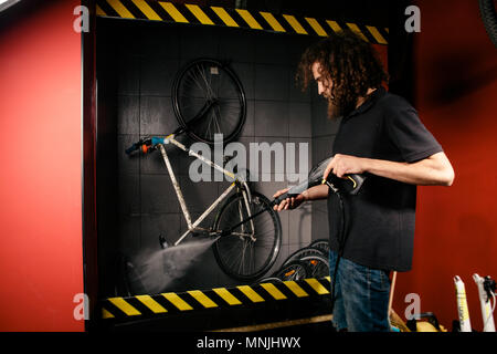 Services professional washing of a bicycle in the workshop. Close-up of hand Young Caucasian stylish man doing bicycle cleaning using automatic electric water pump. Sprays scatter from the pressure Stock Photo