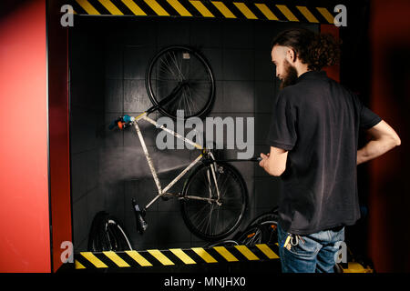 Services professional washing of a bicycle in the workshop. Close-up of hand Young Caucasian stylish man doing bicycle cleaning using automatic electric water pump. Sprays scatter from the pressure Stock Photo