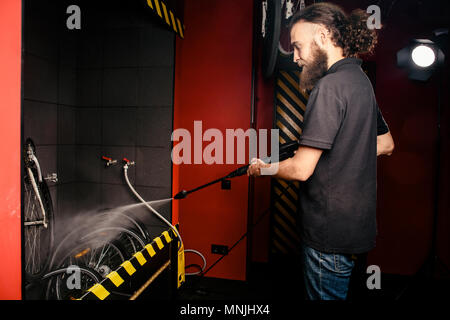 Services professional washing of a bicycle in the workshop. Close-up of hand Young Caucasian stylish man doing bicycle cleaning using automatic electric water pump. Sprays scatter from the pressure Stock Photo