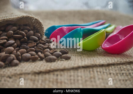 Dark roasted coffee beans on hessian or burlap with colorful plastic kitchen measuring spoons in a shallow dof low angle view Stock Photo